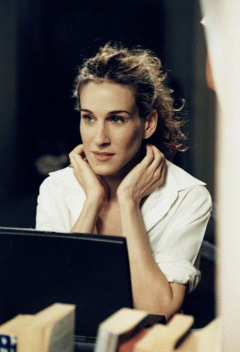 Carrie Bradshaw frante a su computadora usando una blusa blanca y cabello recogido en una coleta alta