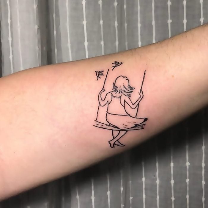 Tatuaje al estilo hand poke de una niña columpiándose en un columpio con un par de aves al rededor