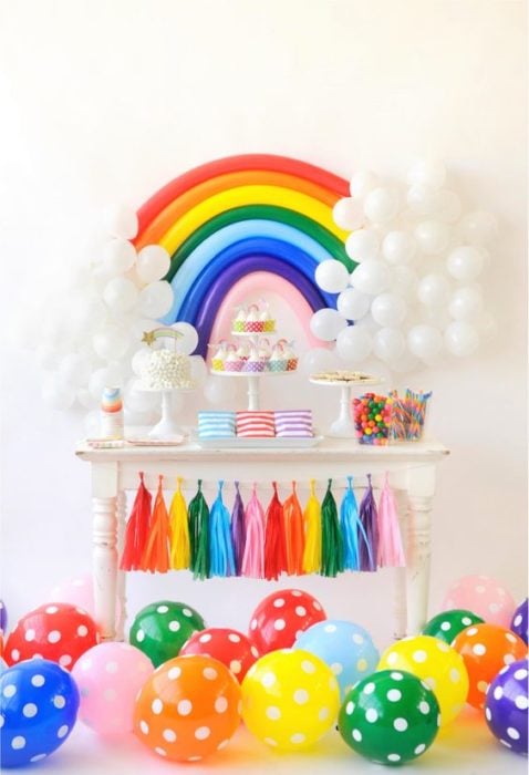 Mesa de dulces adornada con globos de colores con forma de arcoíris y globos blancos como nubes