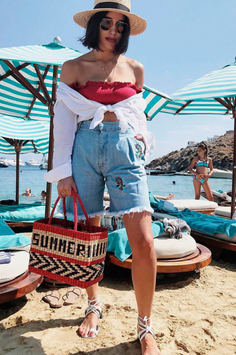 Chica usando bermudas de mezclilla con top de color rojo y camisa de color blanca atada a la cintura 