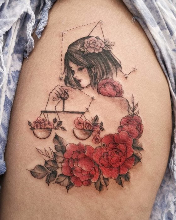 Tatuaje de una chica sosteniendo una balanza y flores de un lado, a tinta negra y de color sobre el área de la pierna