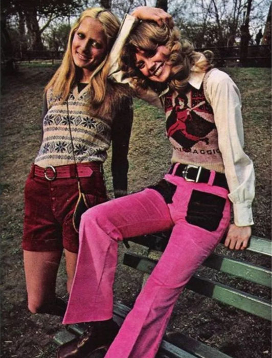 Moda femenina de los 70; amigas sentadas en la banca del parque, suéter de rombos tejido, short de pana rojo, pantalón rosa con negro; peinados y ropa retro