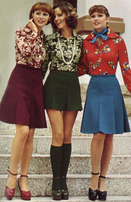 Moda femenina de los 70; tres amigas abrazándose, con blusas floreales guinda, verde y roja, y faldas a la rodilla color vino y azul, zapatillas de tacón con calcetas; ropa y peinados retro