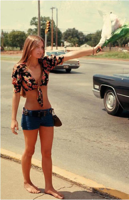 Moda femenina de los 70; mujer pidiendo rait con un ramo de flores, short a la cadera, tio negro floral, descalza; ropa y peinado retro