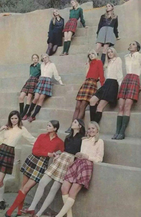 Moda femenina de los 70; grupo de mujeres sentadas en las escaleras con uniforme de estudiante; falda tableada de cuadros, blusas formales blancas, verdes, rojas y negras; ropa y peinados retro