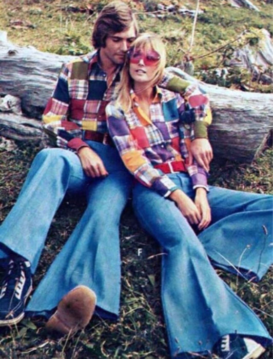 Moda femenina de los 70; pareja sentada en un tronco al aire libre, camisa de retazos de tela, jeans azules acampanados y tenis negros con blanco; ropa retro