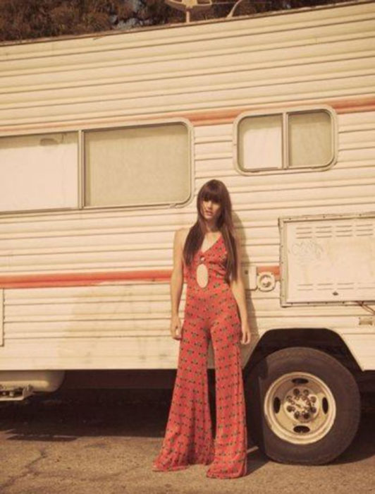 Moda femenina de los 70; mujer frente a un camper con onesie rojo acampanado de flores de escote al ombligo, cabello largo lacio y castaño; ropa retro