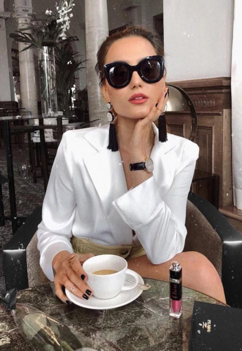 Outfit con blusa blanca; mujer con lentes oscuros, aretes de flecos tomando un café