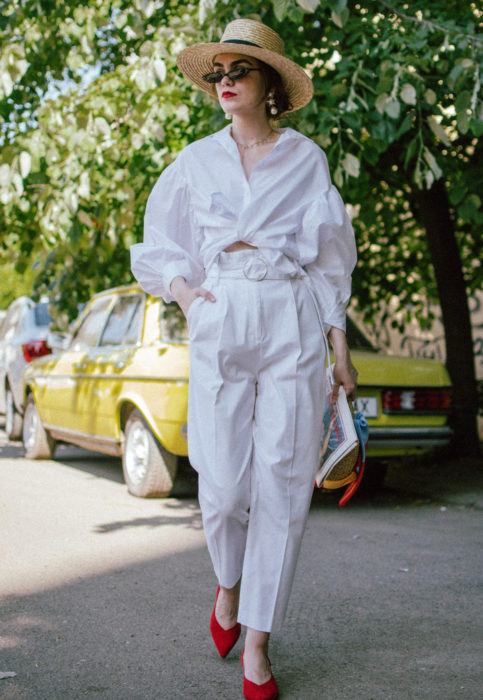 Outfit con blusa blanca; mujer caminando en la calle con sombrero de mimbre, camisa oversized, pantalón blanco y zapatillas rojas