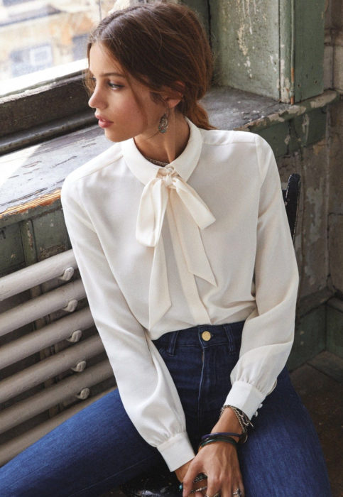 Outfit con blusa blanca; mujer mirando por la ventana con camisa marfil y moño en el cuello