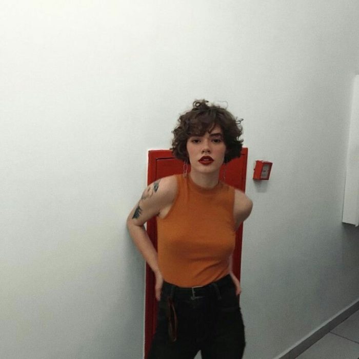 Chica de cabello rizado y muy corto posa con blusa naranja sin mangas y pantalón negro