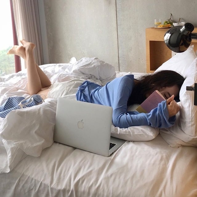 Chica con pijama azul está acostada en la casa con una mac y lee un libro