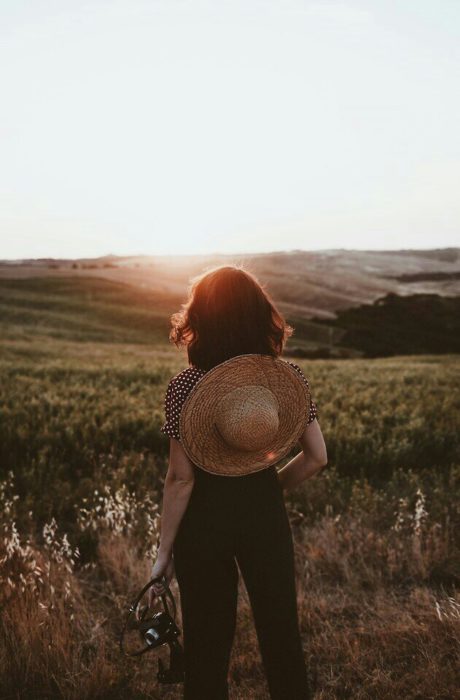 Chica con cabello corto y un sombrero en la espalda mira el atardecer