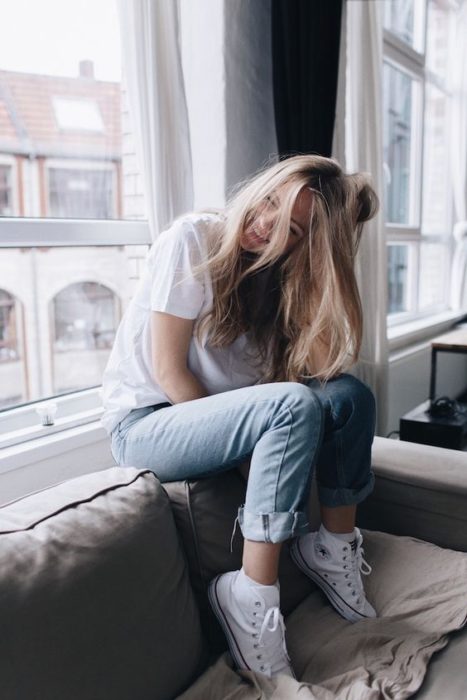 Chica rubia de cabello largo con blusa blanca y jean sentada en un sillón junto a la ventana