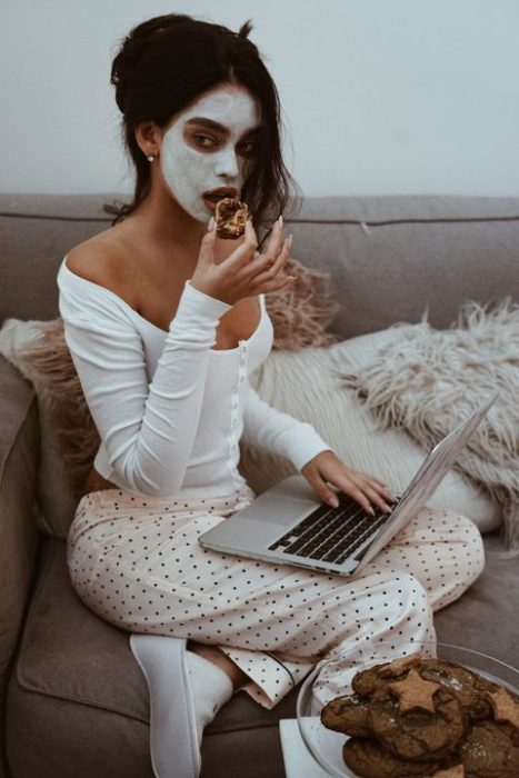 Chica santada en el sillón con su laptop en las piernas usa una mascarilla de arcilla