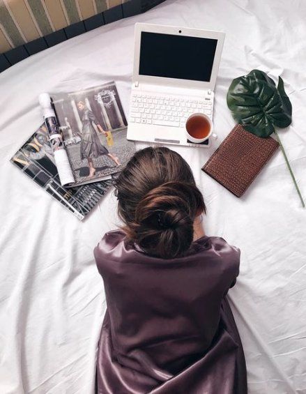 Chica acostada en la cama con revistas, libreta café y una laptop blanca