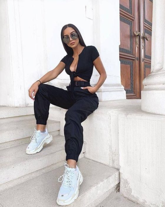 Chica con gafas de sol sentada en escalón viste blusa negra y pantalón negro con tenis blancos