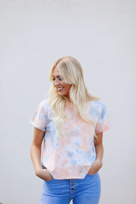 Mujer rubia con cabello suelto posa en una pared blanca con una blusa tie dye en color pastel