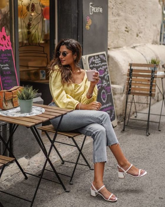 Mujer sentada cruza la pierna y viste blusa amarilla, pantalón gris y tacones blancos cuadrados