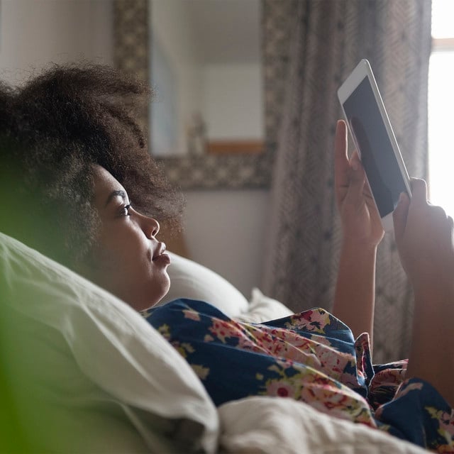 Chica de cabello rizado recostada en su cama leyendo un texto a través de una tablet 