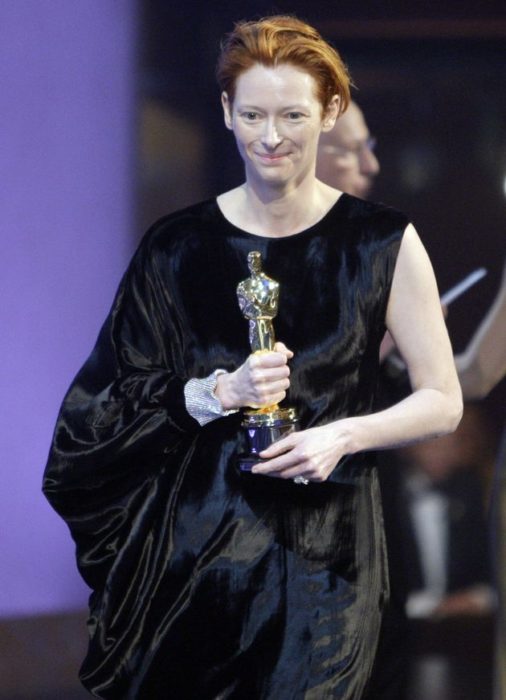 Tilda Swinton con vestido negro de una sola manga sosteniendo una estatuilla de los premios Óscar