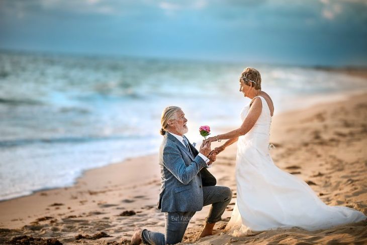 Fotografía por Abigail's Collection & The Groom's Room, pareja de abuelitos regalándose flores a la orilla del mar