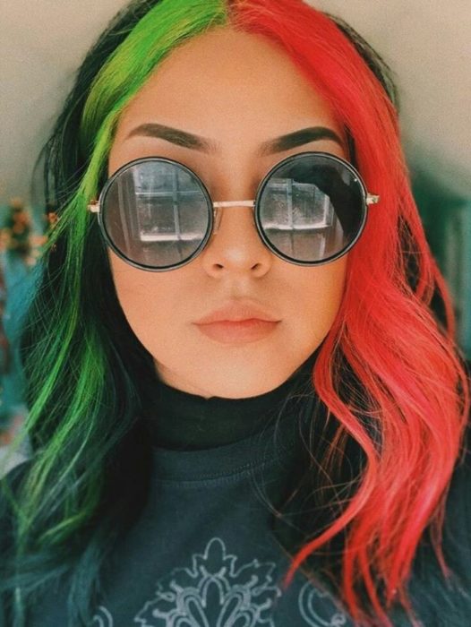 Chica con lentes grandes circulares lleva mechones de cabello verdes y naranjas 