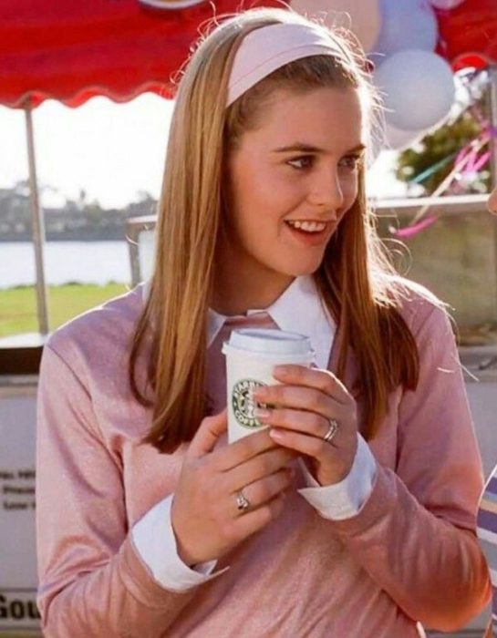 Personaje de Cher en Clueless usando una diadema de color rosa mientras camina y bebe café 