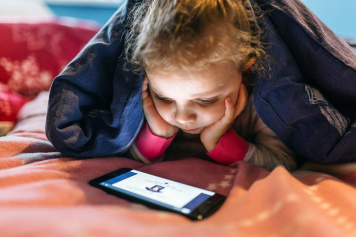 Niña viendo la tablet mientras está bajo las cobijas 