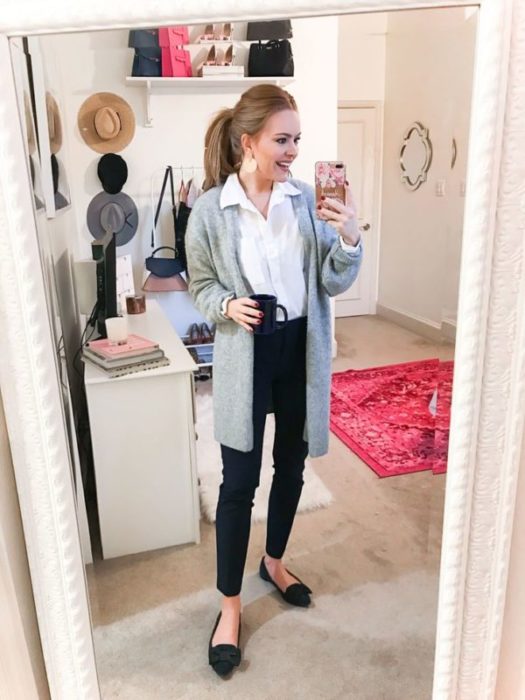 Chica se toma selfie frente al espejo con cardigan gris, blusa blanca y pantalón negro