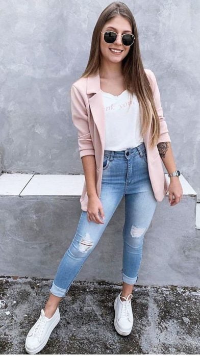 Chica castaña de pelo largo y suelto usa lentes oscuros y viste jeans claros, blusa blanca y saco rosa claro