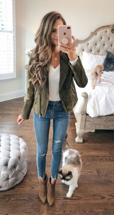 Chica castaña de cabello largo y rizado se toma selfie frente al espejo mientras viste saco verde militar, blusa blanca y jeans