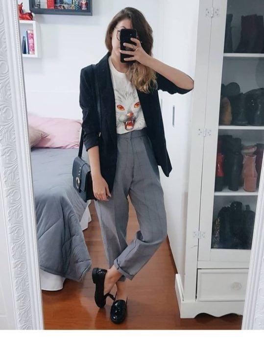 Chica se toma selfie frente al espejo con pantalón gris, saco negro y blusa blanca con estampado de gato