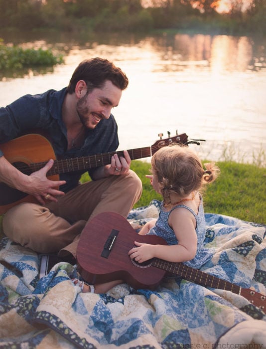 Frases del día del padre; papá tocando la guitarra con si hija en un día de campo al lado de un lago