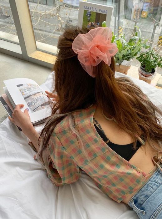 Chica acostada leyendo un libro con media coleta y scrunchie gigante rosa