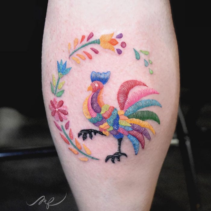 Fernanda Ramírez, tatuadora mexicana crea tatuajes bordados; gallo de colores con flores en el chamorro