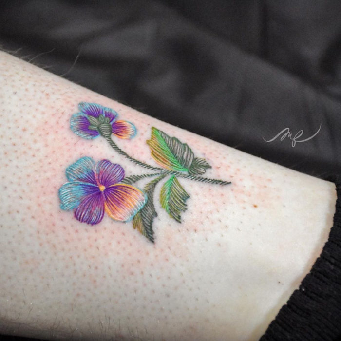 Fernanda Ramírez, tatuadora mexicana crea tatuajes bordados; flores pensamientos morados y azules en la pierna