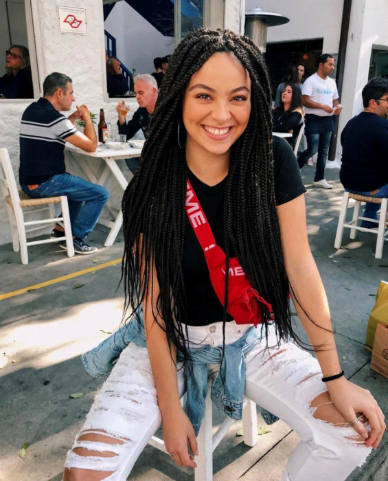 Looks con trenzas africanas; mujer sonriendo sentada en un café, cabello lardo y negro, jeans desgastados