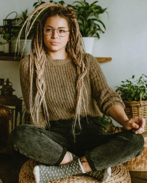 Looks con trenzas africanas; mujer con lentes vintage, suéter tejido café, cabello rubio y largo, sentada en taburete de rafia