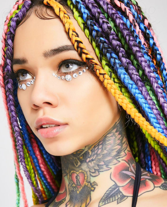 Looks con trenzas africanas; mujer de ojos grises, con cabello de colores arcoíris, anaranjadi, verde, azul, morado y rosa, con tatuaje de búho, rosa y corazón en el cuello, con piedras en los ojos