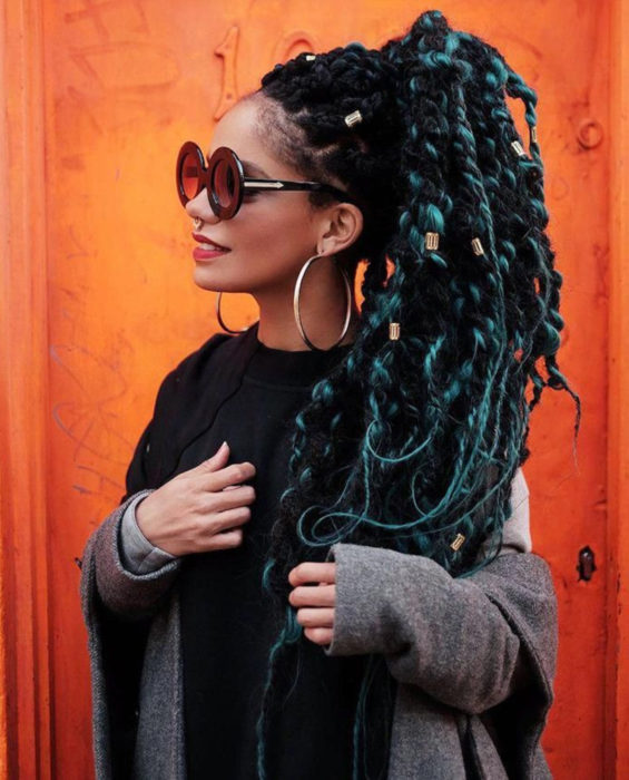 Looks con trenzas africanas; mujer morena con lentes redondos y gruesos, cabello negro y verde largo, peinado en una cola de caballo con accesorios dorados, arracadas grandes y piercing en el tabique