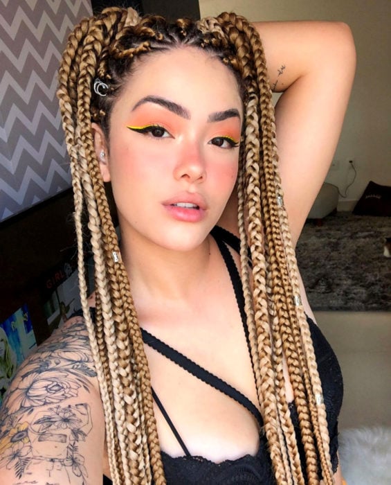 Looks con trenzas africanas; mujer de cabelo largo y rubio con peinado de dos coletas altas, maquillaje natural, tatuaje de flores en el hombro