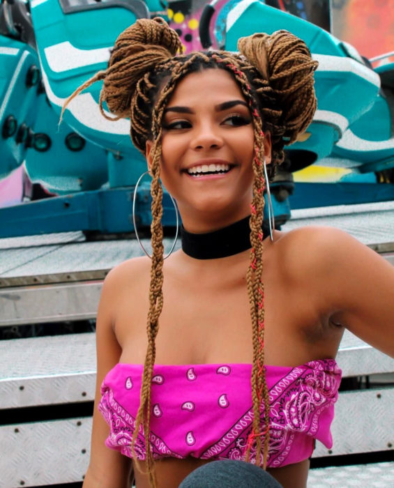 Looks con trenzas africanas; mujer morena sonriendo, con cabello castaño rubio largo, peinado de dos chongos altos, crop top de paliacate rosa y gargantilla negra