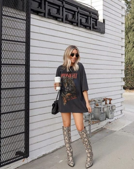 Mujer rubia con melena corta y suelta posa con blusa amplia y larga negra con botas ñargas de animal print