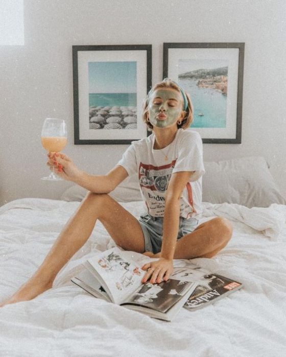 Chica sentada en su cama, bebiendo jugo de naranja y llevando una mascarilla de arcilla verde