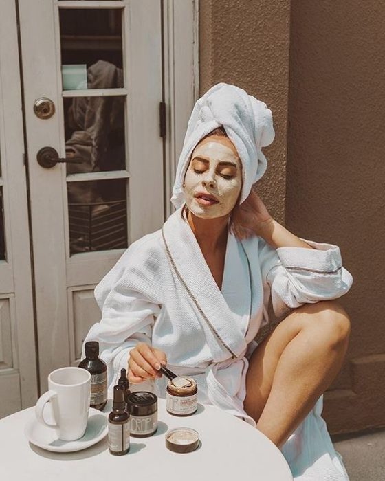 Chic con bata de baño sentada en una silla blanca aplicando una mascarilla en el rostro