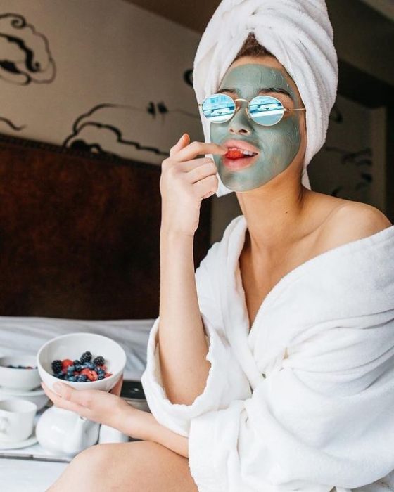 Chica sentada en la orilla de una cama, con una toalla enredada en la cabeza, llevando lentes azules y comiendo frutos rojos