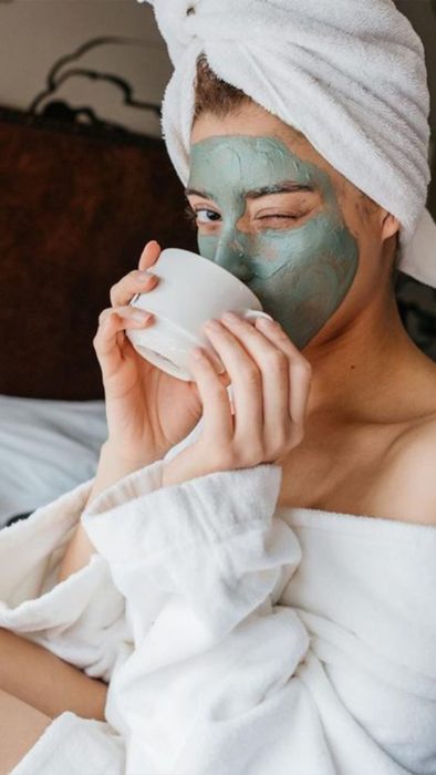 Chica bebiendo té, guiñando el ojo y llevando una mascarilla de arcilla verde