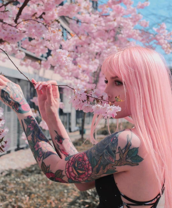 Cabello color rosa cherry blossom, flor de cerezo; mujer con tatuajes en los brazos junto a un árbol de flores rosas, cabello lacio y largo