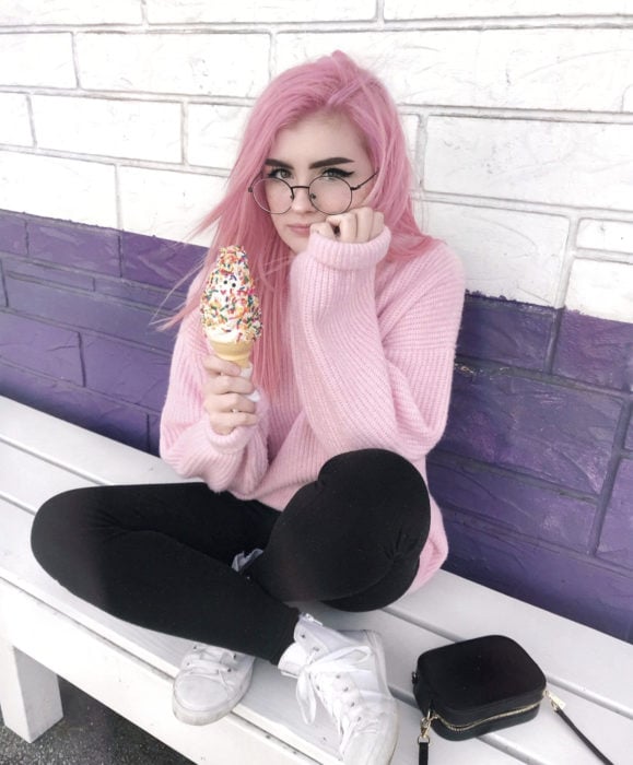 Cabello color rosa cherry blossom, flor de cerezo; chica sentada comiendo un helado con chispas de colores, suéter oversozed tejido, lentes redondos vintahe, cabello lacio y largo teñido
